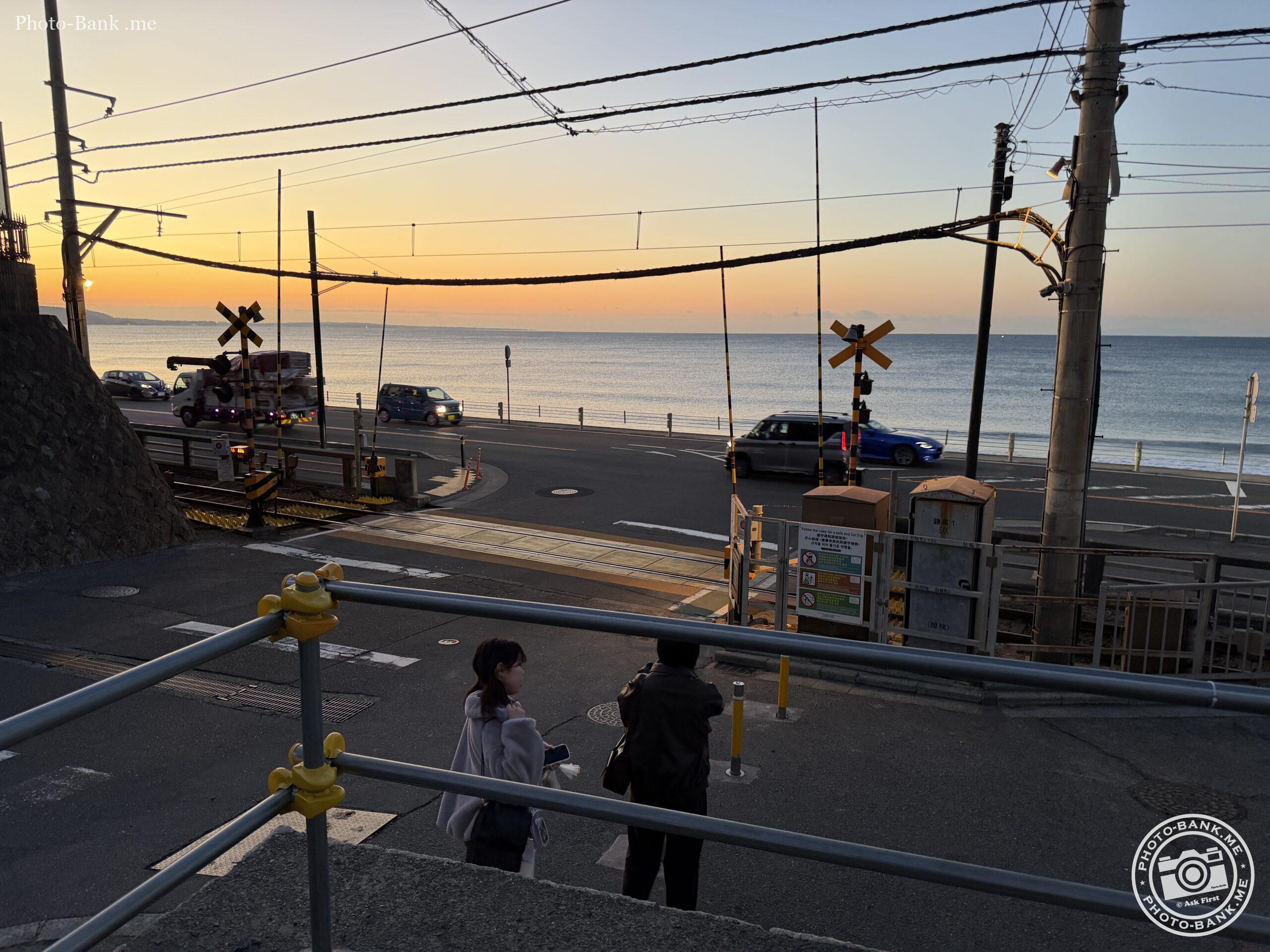 2025年12月撮影、鎌倉高校前駅の夕暮れ時、七里ヶ浜と江の島がシルエットで見える。踏切を渡る人影、道路を走る車。