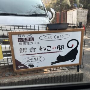 緑の中の古民家風保護猫カフェ「鎌倉ねこの間」の看板と、背景の白い車。