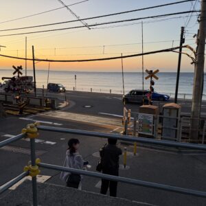 2025年12月撮影、鎌倉高校前駅の夕暮れ時、七里ヶ浜と江の島がシルエットで見える。踏切を渡る人影、道路を走る車。